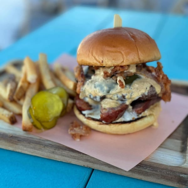 A BBQ burger with sauce, crispy onions, and bacon, served with fries and pickles on a wooden board.