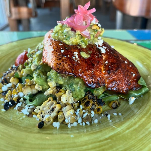 Grilled salmon with guacamole, pickled onions, charred corn, and crumbled cheese on a green plate.
