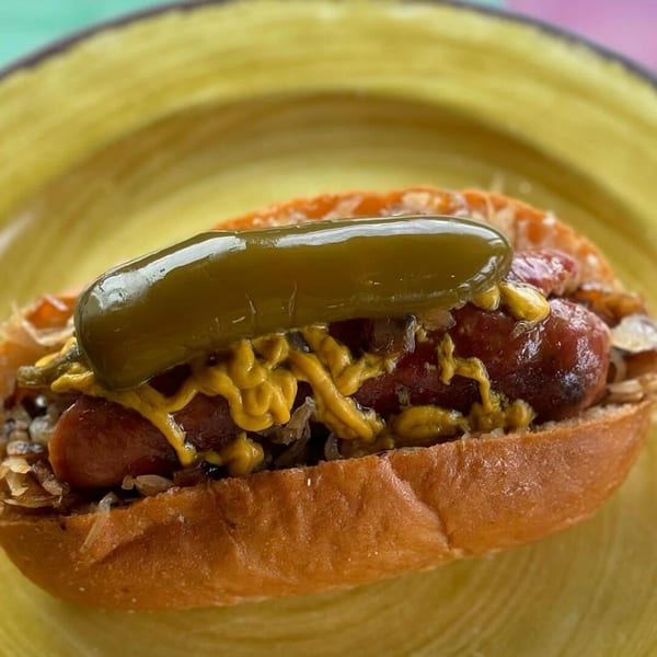 A grilled hot dog in a bun, topped with mustard, onions, and a whole pickled jalapeño.