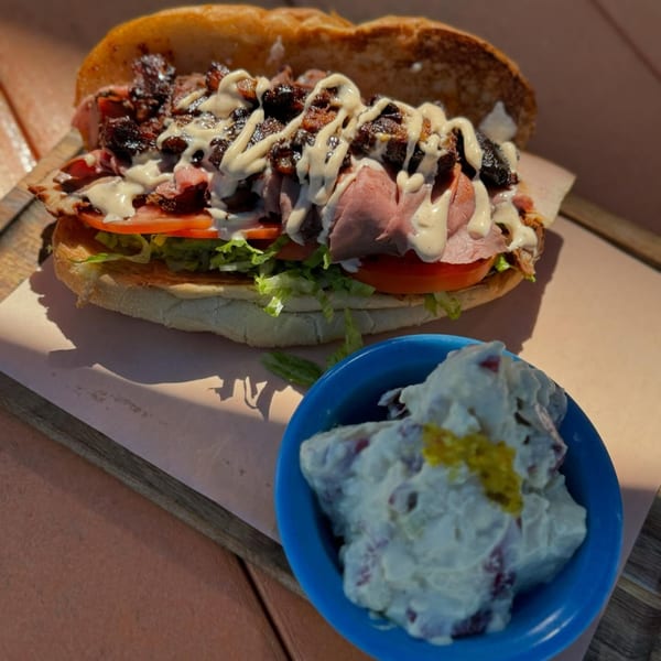 A BBQ sandwich with meat, lettuce, tomato, and sauce, served with a side of potato salad.