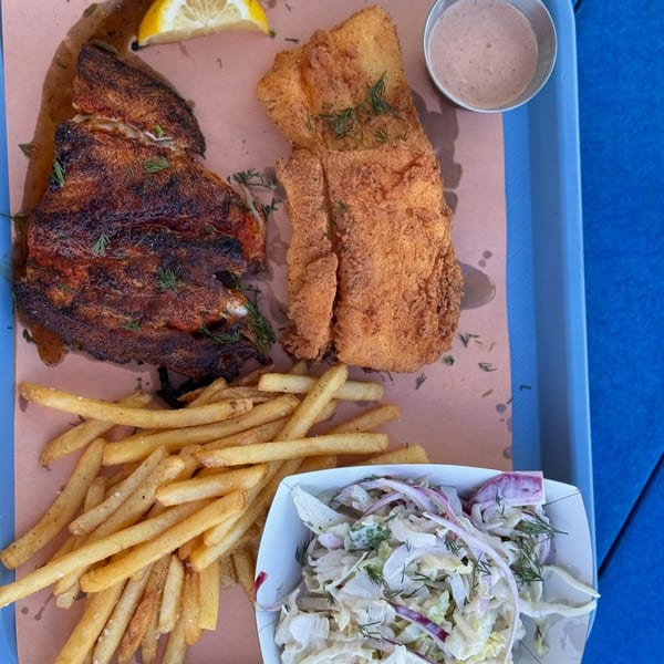A tray with blackened fish, fried fish, french fries, coleslaw, and a side of sauce.