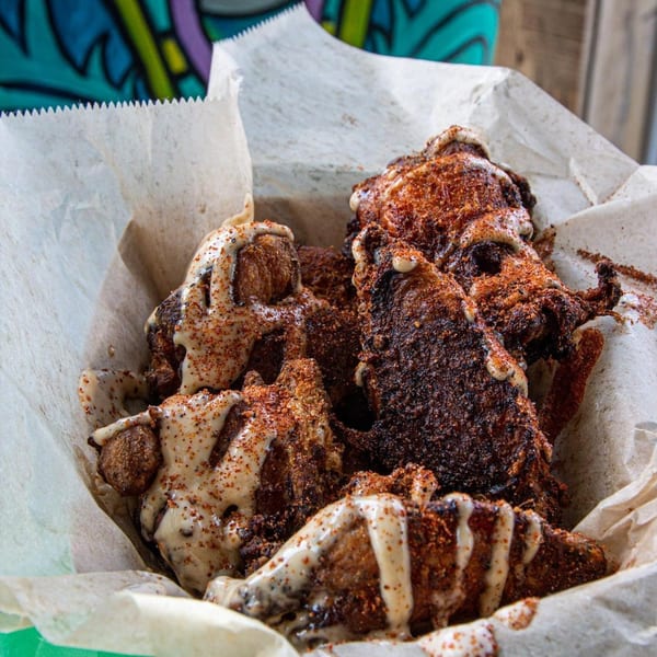 Crispy BBQ chicken wings, drizzled with white sauce and sprinkled with red seasoning, in a paper-lined basket.