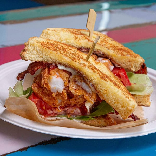 Two grilled sandwiches with fried fish, bacon, lettuce, tomato, and sauce on a white plate.